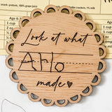 A wooden fridge magnet with the text 'Look at what Remi made' engraved on it, displayed on a refrigerator with a colorful artwork in the background.
