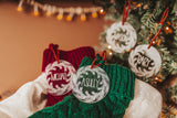 Clear acrylic stocking tags laser engraved with an evergreen wreath and a first name.  They are tied with a suede cord and sitting on top of festive red and green stockings.