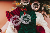 Clear acrylic stocking tags laser engraved with an evergreen wreath and a first name.  They are tied with a suede cord and sitting on top of festive red and green stockings.