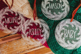 Clear acrylic stocking tags laser engraved with an evergreen wreath and a first name.  They are tied with a suede cord and sitting on top of festive red and green stockings.