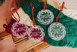 Clear acrylic stocking tags laser engraved with an evergreen wreath and a first name.  They are tied with a suede cord and sitting on top of festive red and green stockings.