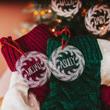 Clear acrylic stocking tags laser engraved with an evergreen wreath and a first name.  They are tied with a suede cord and sitting on top of festive red and green stockings.