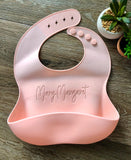 Pink Silicone toddler Bib with food catching pocket "personalized" laser engraved with child's name in cursive script on a wood grain background.