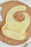 Yellow Silicone toddler Bib with food catching pocket "personalized" laser engraved with child's name in cursive script displayed on a nature fiber round placemat.