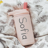 Pink color 12-ounce stainless steel, flip-top straw tumbler. The tumbler is laser engraved and personalized with the name of the child, the font is a simple handwritten style. The tumbler is sitting on a white and black patterned fabric.