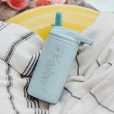 Mint color 12-ounce stainless steel, flip-top straw tumbler. The tumbler is personalized with a dinosaur that its body is the name of the child. The tumbler is nestled on a beach towel.