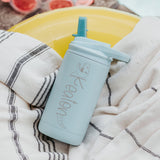 Mint color 12-ounce stainless steel, flip-top straw tumbler. The tumbler is personalized with a dinosaur that its body is the name of the child. The tumbler is nestled on a beach towel.