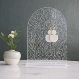 Clear acrylic earing holder with an arch shape and floral designs engraved into the glass.  There are two pairs of earrings hanging on the organizer