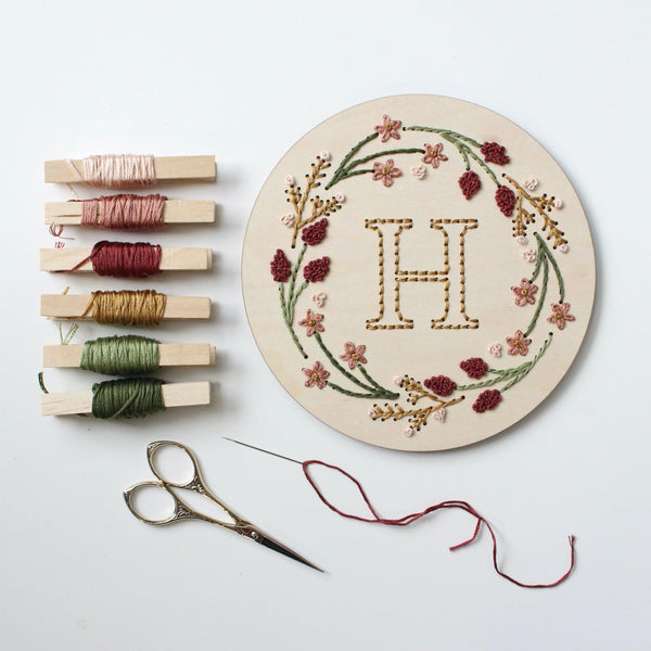 A flat lay of a hand embroidery project featuring a 6-inch round wooden embroidery blank with a floral design and the letter "H" stitched in the center. The floral wreath is embroidered in pink, burgundy, mustard yellow, and green thread. To the left are six clothespins wrapped with coordinating thread colors, a pair of embroidery scissors, and a needle threaded with burgundy floss.