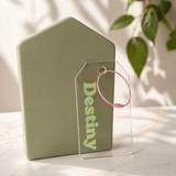 Green house-shaped product with clear acrylic 'Destiny' luggage tag and pink ring on marble surface