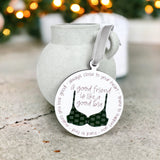 A white ornament with a green, patterned bra that states "a good friend is like a good bra. Always close to your heart, there to support you, hard to find, makes you look good" on a cement table with Christmas decor in the background