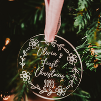 Clear acrylic Christmas ornament with OIivia's first Christmas laser engraved into it with a pink chenille ribbon hanging on a xmas tree