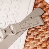 A close-up of a gray silicone watch band compatible with a Fitbit, featuring a personalized engraving that reads 'I have always loved you and always will'. The band is displayed atop a woven mat, with a corner of a white keyboard with round keys visible in the upper left. The intimate engraving provides a sentimental touch to the functional accessory.