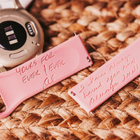 A close-up of a coral-pink watch band with an engraved message 'Yours for ever & ever & ever' lying on a woven straw mat. Next to the band are office supplies: a white keyboard in the top left corner, a polka-dotted pen, and a few gold paper clips. The items are arranged in a casual yet aesthetically pleasing manner, suggesting a creative or personalized workspace.