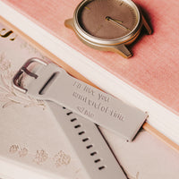 A close-up image showing a pale gray watch band with a personal engraving that reads 'I'll love you until end of time <3 Mo'. The band is displayed over an open notebook with a coral cover, with a blurred spiral notebook in the background. The sentimental note, elegant script, and warm tones convey a sense of intimacy and affection.