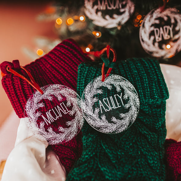 Clear acrylic stocking tags laser engraved with an evergreen wreath and a first name.  They are tied with a suede cord and sitting on top of festive red and green stockings.  