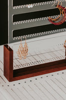 Close up of a clear acrylic freestanding earring holder with a rich wooden base. Displayed with three pairs of earrings on a green background with decorative accents.