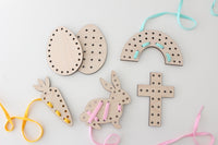 A set of wooden Easter lacing cards for kids on a white background. The set includes two egg-shaped cards with multiple holes around the edges, two rabbit-shaped cards with holes along the body and ears, a cross-shaped card with evenly spaced holes, and a half-circle card resembling a rainbow with holes following the arc. Each card has colorful shoelace-like strings, in yellow, pink, and light blue, threaded through some of the holes, designed to help develop fine motor skills and hand-eye coordination.