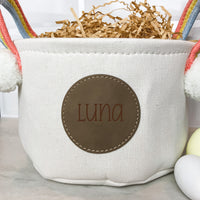 Close up of White Easter Basket with Rainbow Handles with Round Leather patch with the name Luna engraved on it. Two Easter eggs are in the foreground