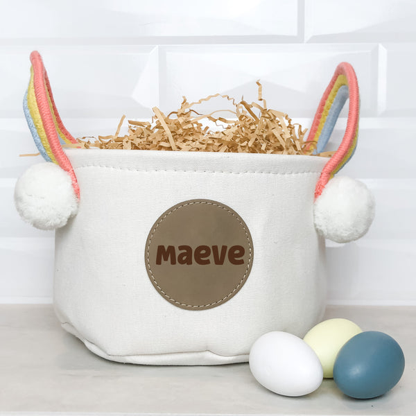 White Easter Basket with Rainbow Handles with Round Leather patch with the name Maeve engraved on it.  Three Easter eggs are in the foreground