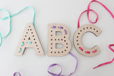 Wooden alphabet lacing cards in the shape of uppercase letters 'A', 'B', and 'C' on a white background. Each letter has multiple lacing holes along its contour. The 'A' has a light blue lace partially threaded through, the 'B' has a purple lace, and the 'C' has a pink lace. The colorful laces lie loosely around the letters, creating a playful and educational activity for children to develop fine motor skills and letter recognition.