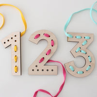 Wooden number lacing cards for kids, featuring the numbers '1', '2', and '3' on a white background. Each number has several holes for lacing, with colorful shoelace-like strings threaded through some of them: yellow for '1', pink for '2', and light blue for '3'. These educational toys are designed to enhance children's fine motor skills and number recognition through a fun, hands-on activity.