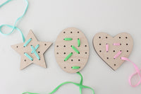 Three wooden lacing shapes laid out on a white background: a star with light blue lace, an oval with green lace, and a heart with pink lace. The laces are threaded through some of the holes in each shape, illustrating a fun and educational activity designed to enhance hand-eye coordination and fine motor skills for children.