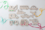 An assortment of wooden lacing cards shaped like various modes of transportation on a white background. The collection includes a car, ambulance, fire truck, helicopter, airplane, bus, and a boat, each with holes along their outlines for lacing. Colorful laces in light blue, yellow, and pink are threaded through some of the cards, creating a fun and engaging activity for kids to develop fine motor skills and learn about different vehicles.