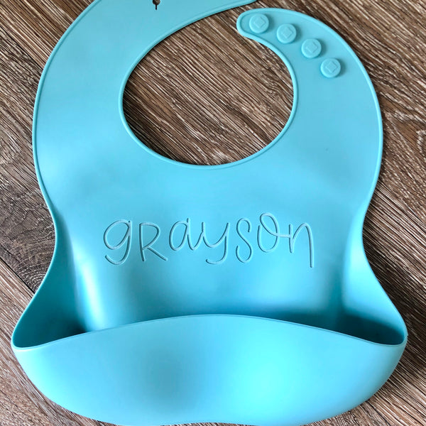 Blue Silicone bib with Grayson engraved in mixed case font on wooden background