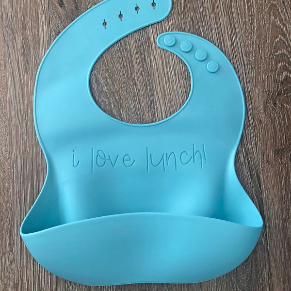 Blue soft silicone baby bib, with a food catching pocket. Laser engraved with "I Love Lunch!" in a simple handwritten syle font. Bib is on a wood grain background.