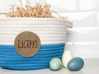 Blue and white braided Easter basket with a leather patch engraved with the name Liam with kraft basket filler and 3 colored eggs in the foreground
