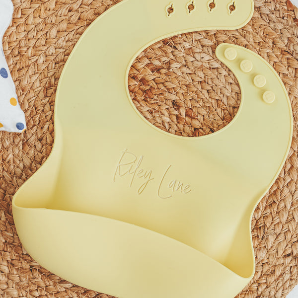 Yellow silicone baby bib with the name Riley Lane laser engraved on a textured background by Legacy and Light