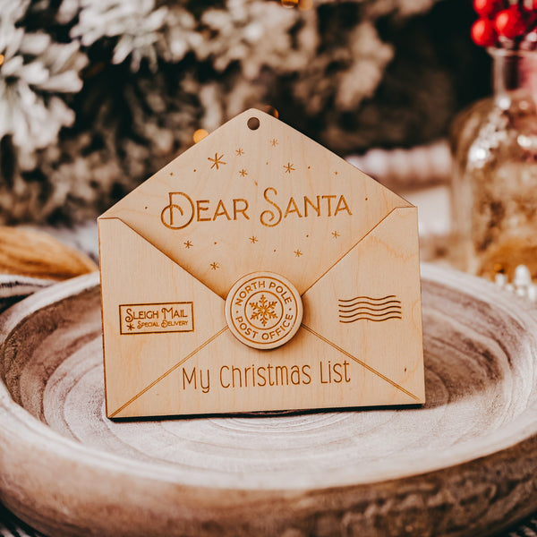Wooden Christmas Ornament shaped as an open envelope. The flap of the envelope has "Dear Santa" with stars around. The left hand side has "SLEIGH MAIL Special Delivery" The bottom has "My Christmas List" In the middle it has a circle with "north pole post office" 