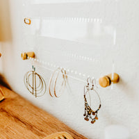 Close up image of Clear Acrylic Earring holder hung on a white textured wall with several pairs of earrings displayed and Gold hardware at each corner by Legacy and Light 