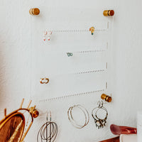 Clear Acrylic Earring holder hung on a white textured wall with several pairs of earrings displayed and Gold hardware at each corner