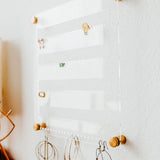 Clear Acrylic Earring holder hung on a white textured wall with several pairs of earrings displayed and Gold hardware at each corner by Legacy and Light