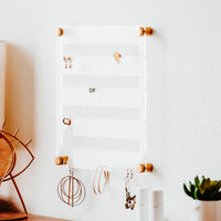 Clear Acrylic Earring holder hung on a white textured wall with several pairs of earrings displayed and Gold hardware at each corner by Legacy and Light