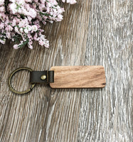 Image shows the back of the wood keychain (no engraving) and shows detail of the vegan leather strap and the brass type fastner. The keychain is sitting on a wood grain background with flower accents.