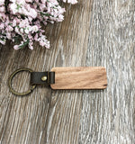 Image shows the back of the wood keychain (no engraving) and shows detail of the vegan leather strap and the brass type fastner. The keychain is sitting on a wood grain background with flower accents.