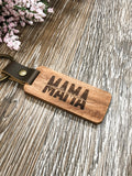 Image shows a laser engraved wood keychain with the word "Mama" and two names over the top in a smaller font. The brass color keyring is attached by a dark brown vegan leather strap that is secured with a brass type fastner. The keychain is on a wood grain background.