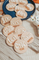 A collection of wooden milestone discs spread out on a textured beige fabric. Each disc has a different milestone from 'one MONTH' to 'one YEAR' written in raised cursive white lettering. The discs are arranged in ascending order, capturing the monthly growth of a newborn baby. In the background, there’s a blue plate and a plush toy, adding to the warm, celebratory feel of the scene.