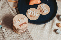 A stack of wooden milestone discs for a newborn baby, with the topmost disc reading 'eleven MONTHS' in raised cursive white lettering. Beside the stack are two individual discs with 'hey y'all' and 'one YEAR'. These discs are arranged on a textured beige fabric, with a navy blue hat in the background, creating a charming and celebratory setting for commemorating baby's monthly growth milestones.