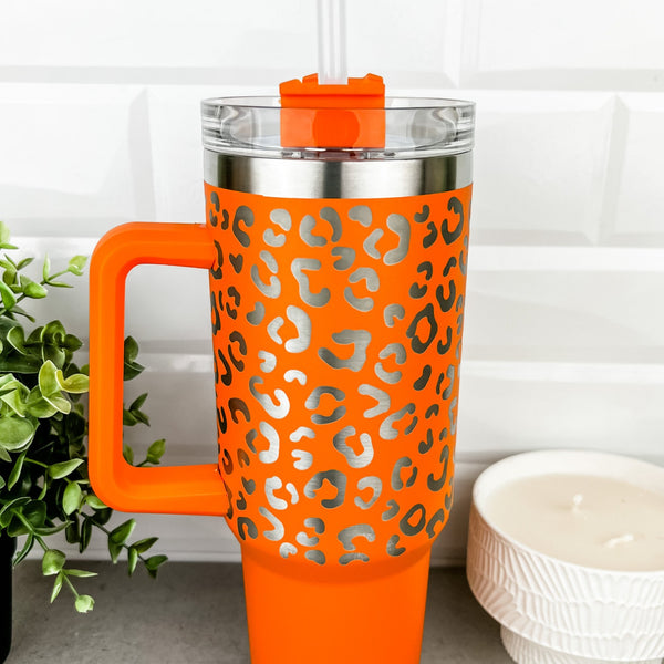 A bright orange tumbler with leopard print engraved across the top half.  The tumbler has a clear lid and 12 in straw.  It has a handle and is similar in design to the Stanley Quencher 40 oz Tumblers.  