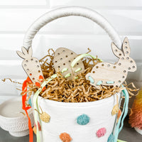 A festive Easter basket with a white fabric exterior decorated with multicolored polka dots, filled with shredded brown paper. Two wooden lacing cards, one shaped like a carrot with an orange lace and the other like a bunny with a light blue lace, protrude from the basket. The basket is placed on a gray surface, with a soft shadow indicating a bright setting, creating a cheerful, holiday-themed scene.