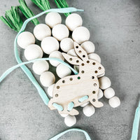 A wooden bunny-shaped lacing card with holes for threading on a gray surface, with a light blue lace partially threaded through the holes. Scattered around are white wooden beads and strands of artificial green chives, adding to the playful and crafty atmosphere suggestive of a child's educational activity or a festive Easter decoration.