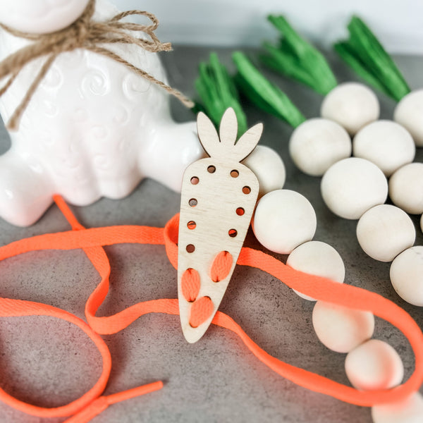 A wooden carrot-shaped lacing card with holes along its length and an orange lace threaded through some of the holes, placed on a gray countertop. In the background, there's a ceramic bunny with a jute twine bow, a bunch of artificial green chives, and a pile of white wooden beads, contributing to a playful and crafty Easter-themed setting.
