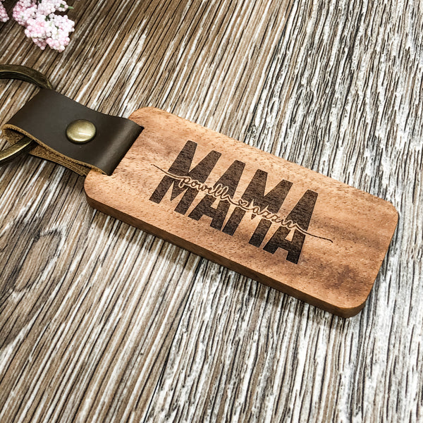 laser engraved wood keychain with the word "Mama" and two names inset. The brass color keyring is attached by a dark brown vegan leather strap that is secured with a brass type fastner. The keychain is on a wood grain background.