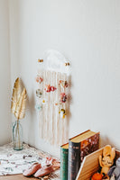 Another view of the hair bow holder hung. on a white wall with hair bows on the cotton strings. On a white background with books and gold flowers as accents.