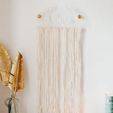 Image is of an arylic hair bow holder. it is a half circle, clear acrylic, with white acrylic leaf accents and personalized name on the front. Cotton string hangs from the bottom of the half circle. The hair bow holder is hing on a white wall by two round gold wall brackets.
