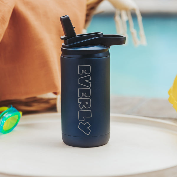 Midnight Blue color 12-ounce stainless steel, flip-top straw tumbler. The tumbler is laser engraved and personalized with the name of the child, the font is in the block style. The tumbler is sitting on a white table with a yellow pool floaty in the background.